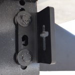 Close-up of a black metal mounting plate with two large bolts on a textured steel beam, showing a vertical slotted hole.