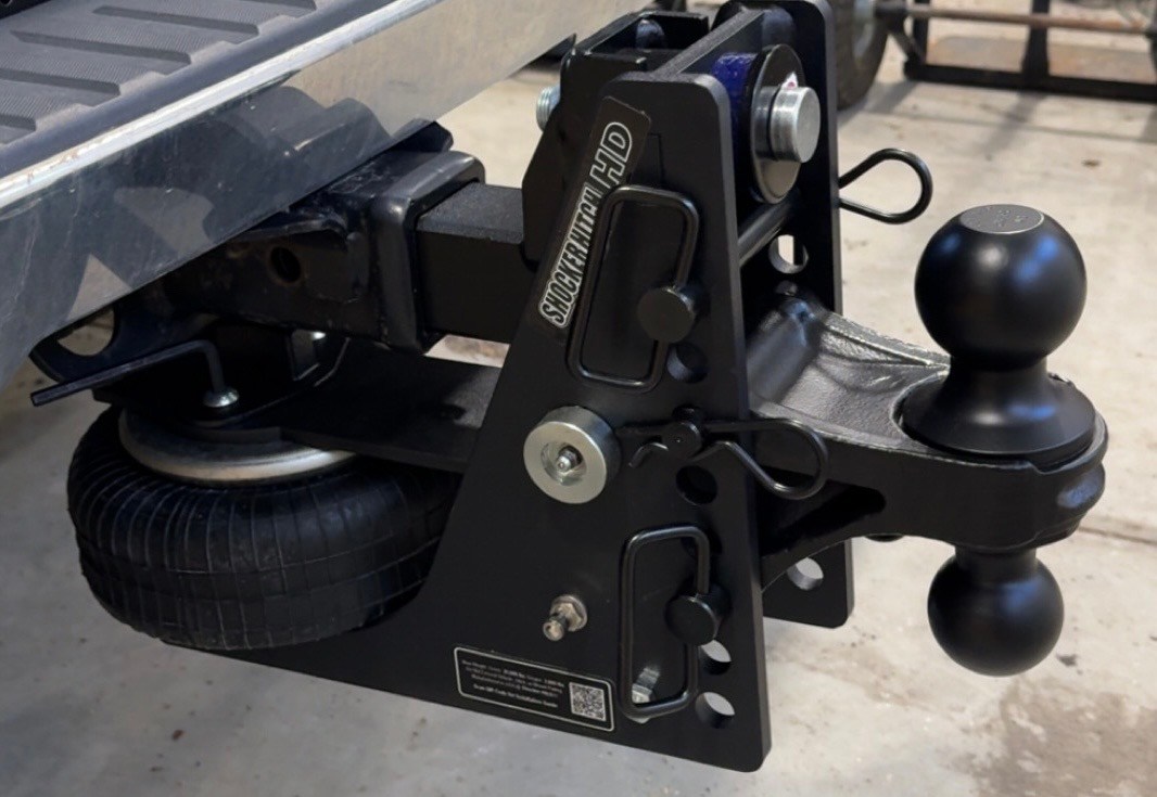 Close-up of a heavy-duty trailer hitch assembly showing a black steel frame, locking pin, and a rounded hitch ball with a spare tire visible underneath.