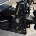 Close-up of a heavy-duty trailer hitch assembly showing a black steel frame, locking pin, and a rounded hitch ball with a spare tire visible underneath.
